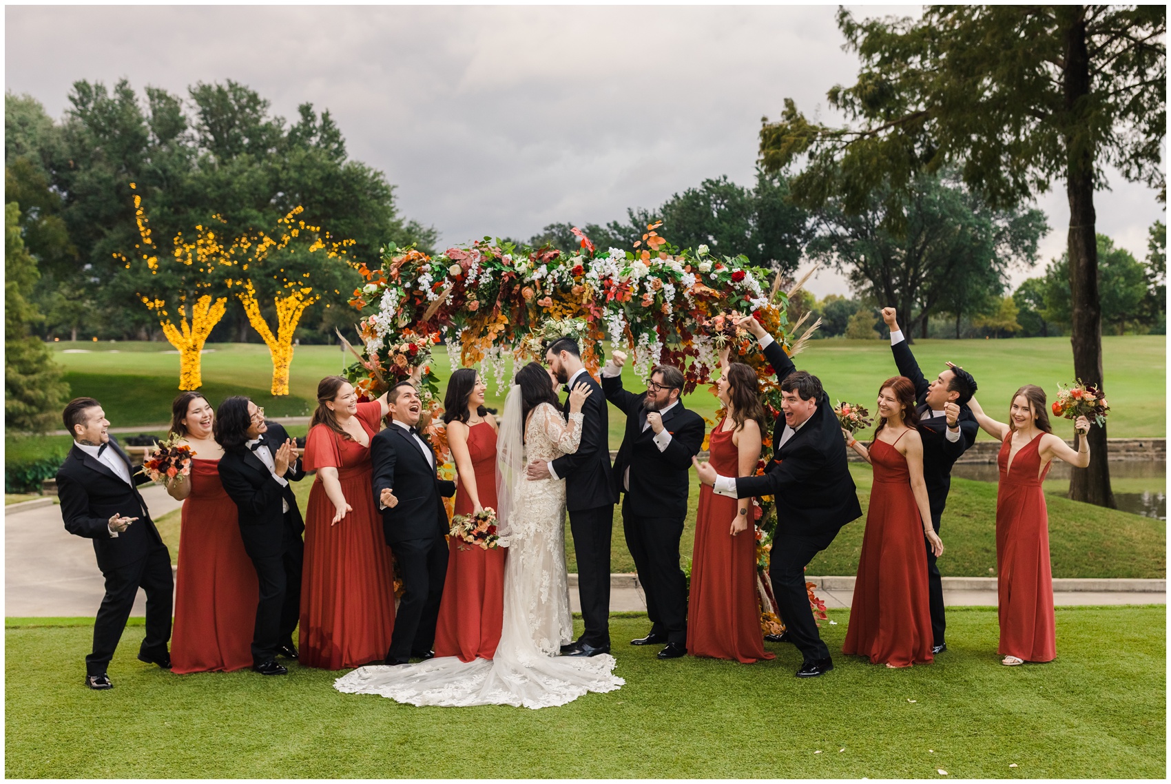 A bride and groom kiss under a colorful floral arbor with bridal party cheering around them