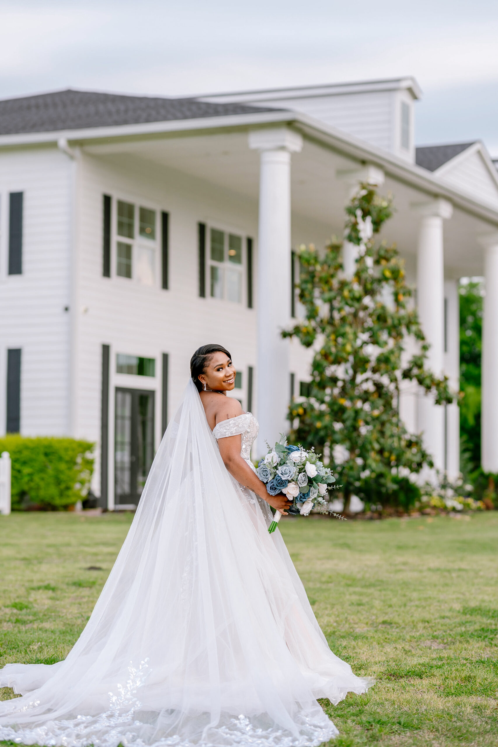 A bride with her wedding dress and long veil standing in front of The Pearl at Sabine Creek