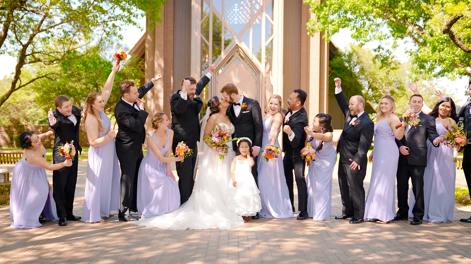 A bridal party posing in front of The Marty Leonard Chapel and cheering as a newlyweds share a kiss