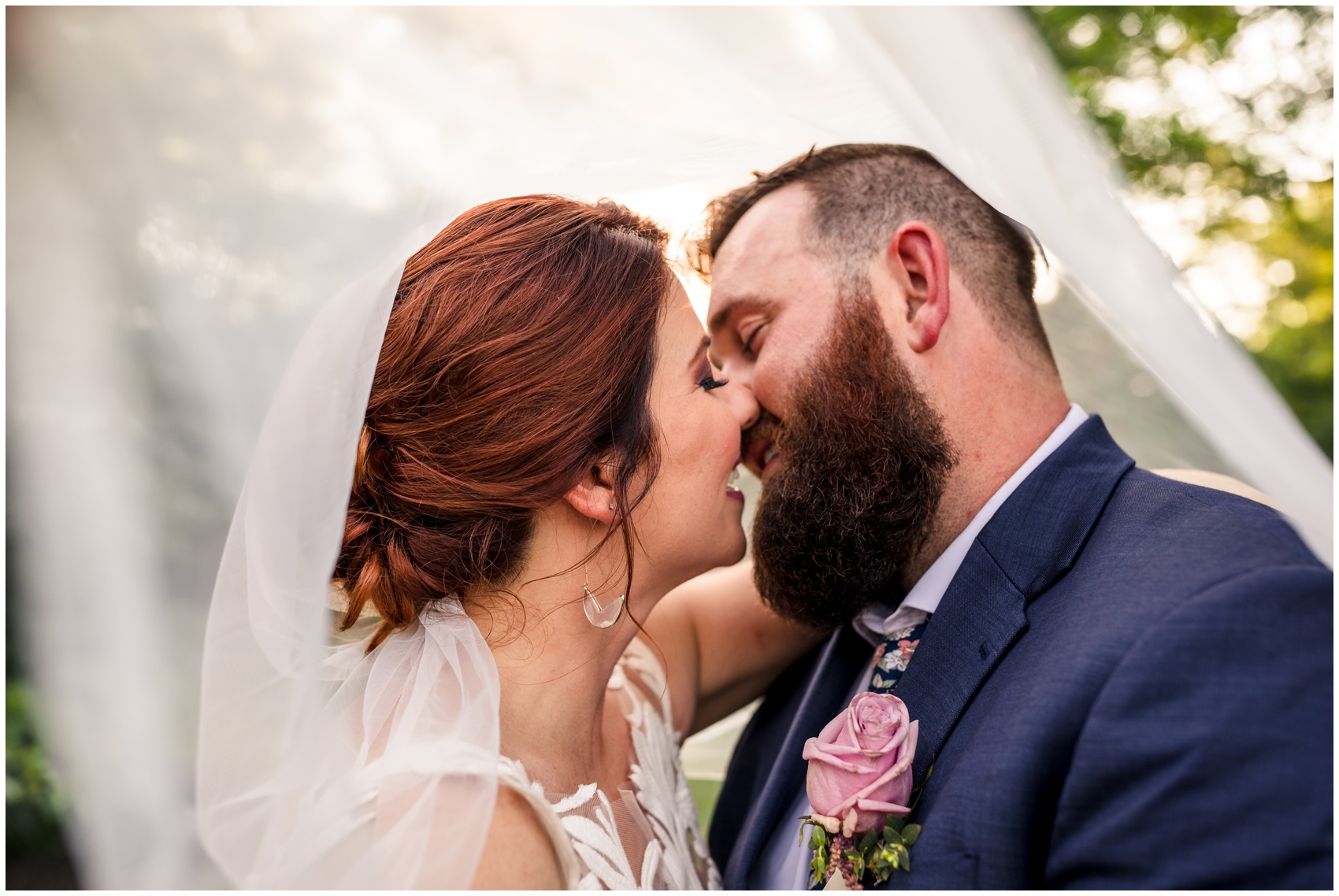 Newlyweds lean in for a happy kiss under the veil at sunset during their D'Vine Grace Vineyard in a blue suit and lace dress