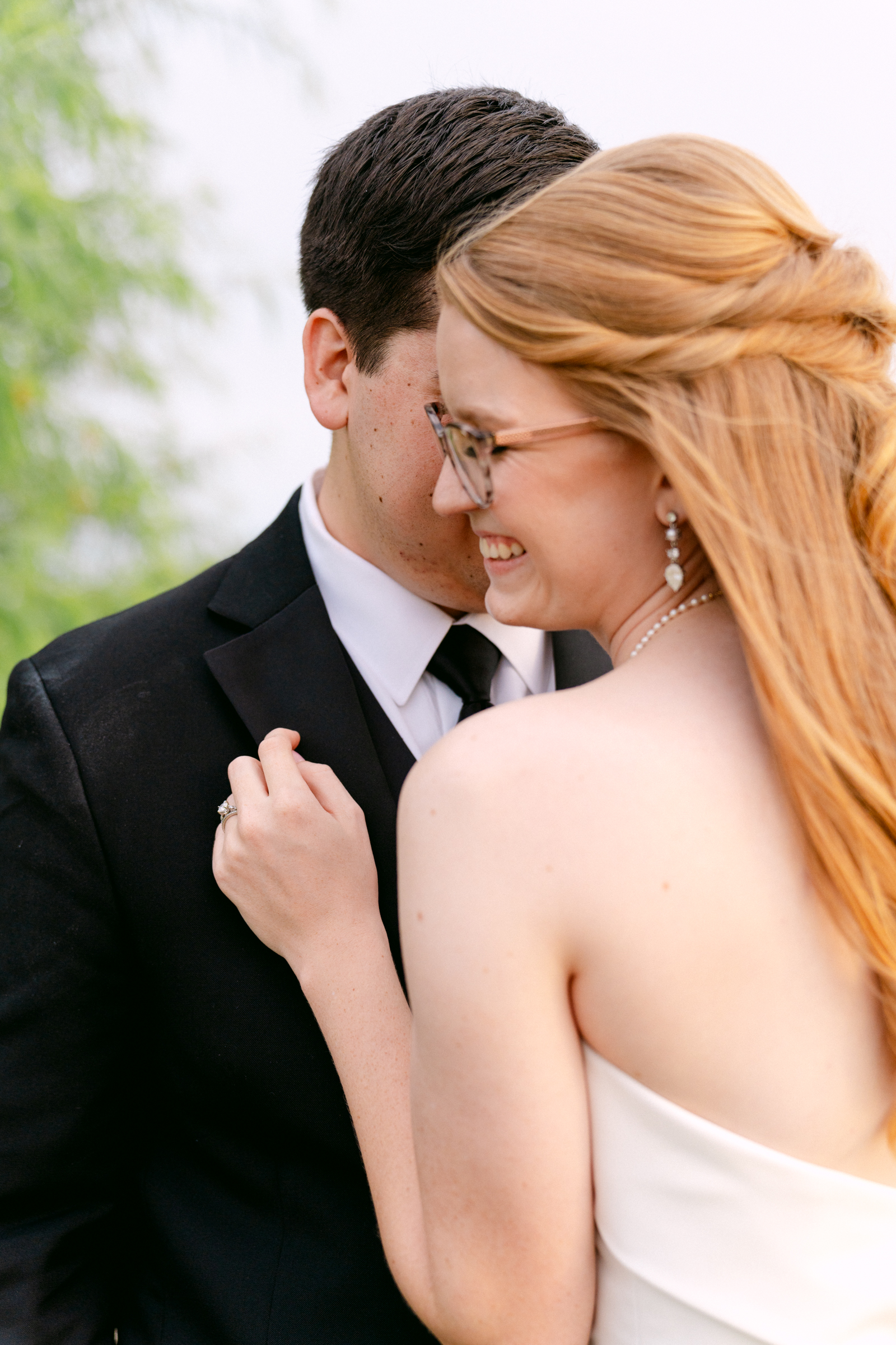 A bride and groom giggle and snuggle while standing under a tree at sunset