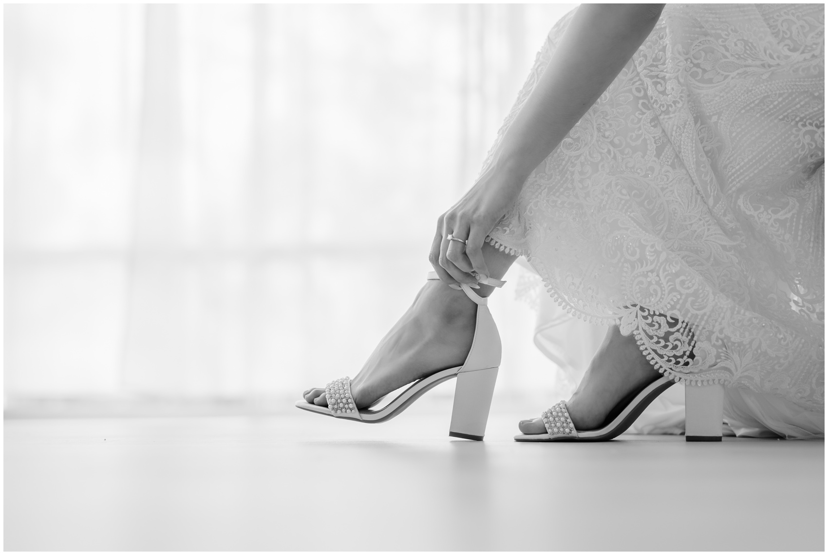 Details of a bride putting on her shoes in a lace dress while sitting in a large window at her Hillside Estate Wedding