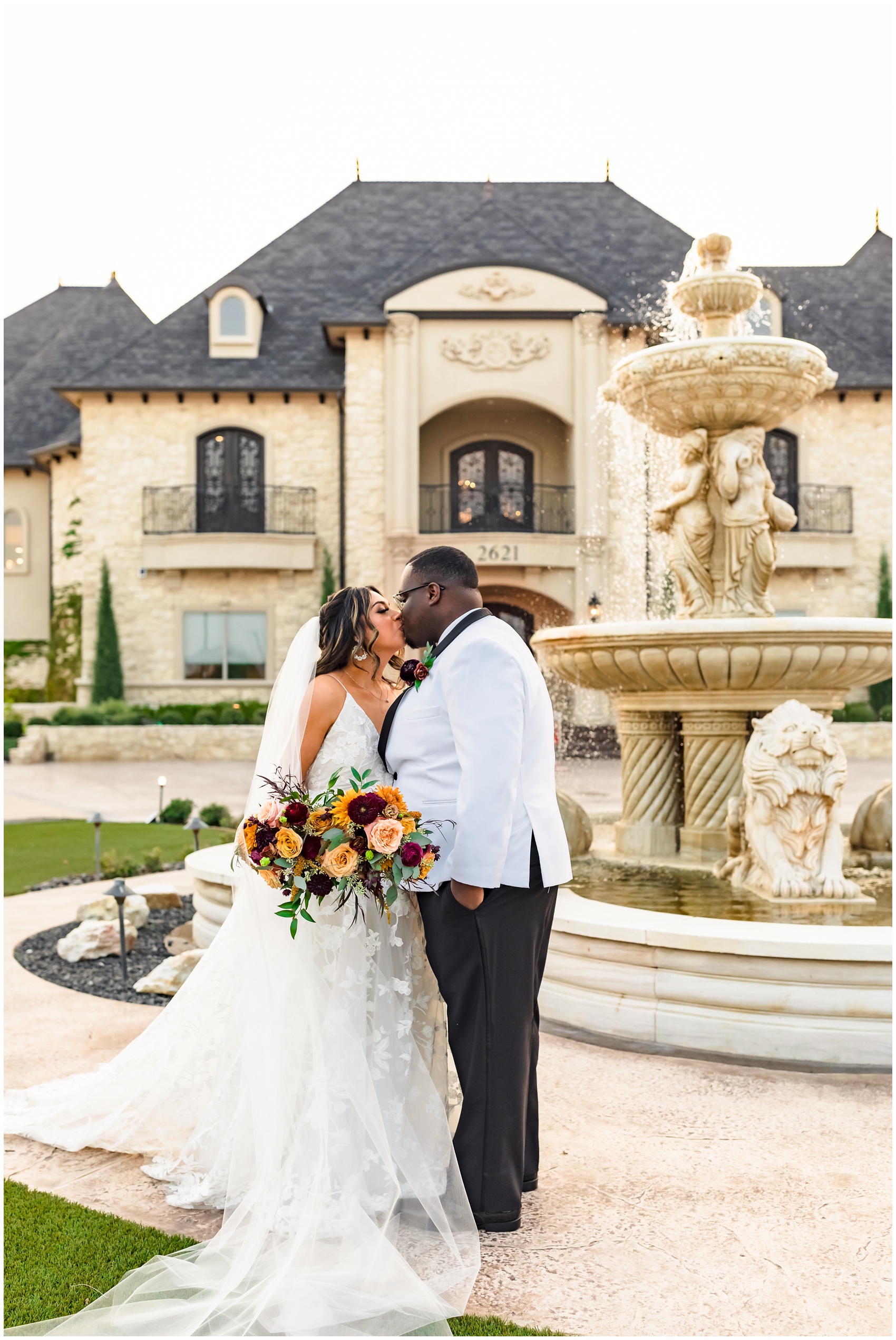 Newlyweds kiss by the garden fountain in a white tux jacket and long flowing lace dress