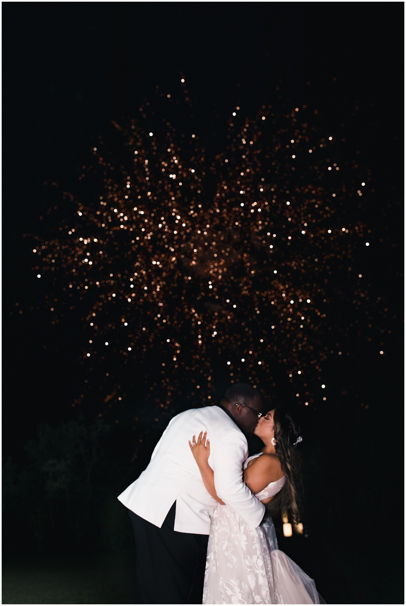 Newlyweds kiss under fireworks in a white tuxedo and lace dress