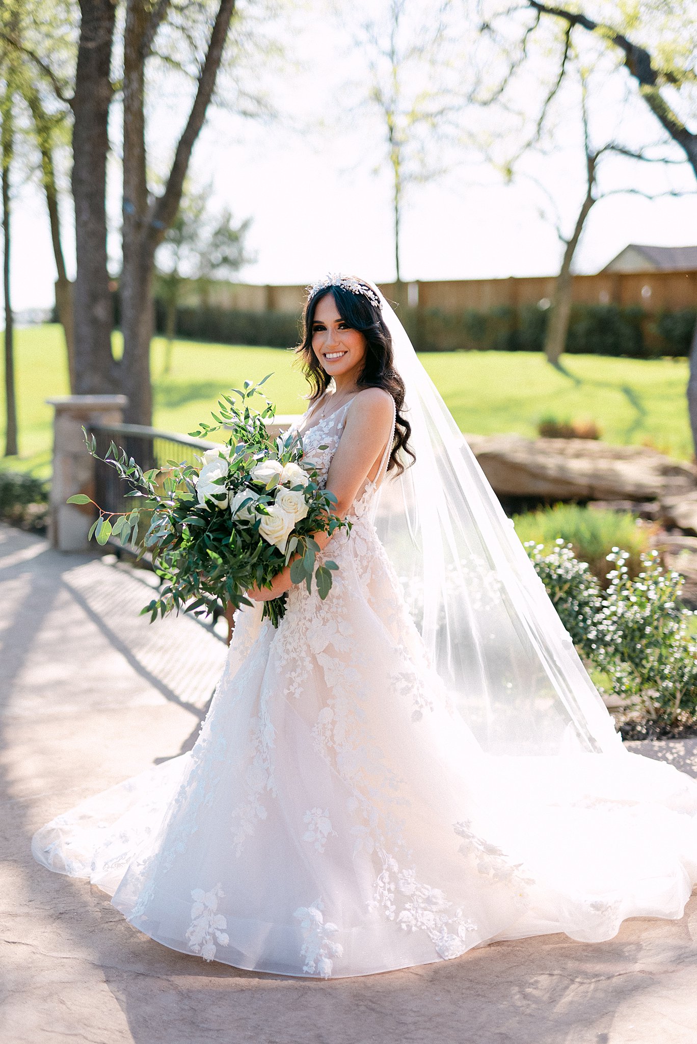 A bride smiles while standing in a garden sidewalk holding her white rose bouquet at her Lone Star Mansion Wedding