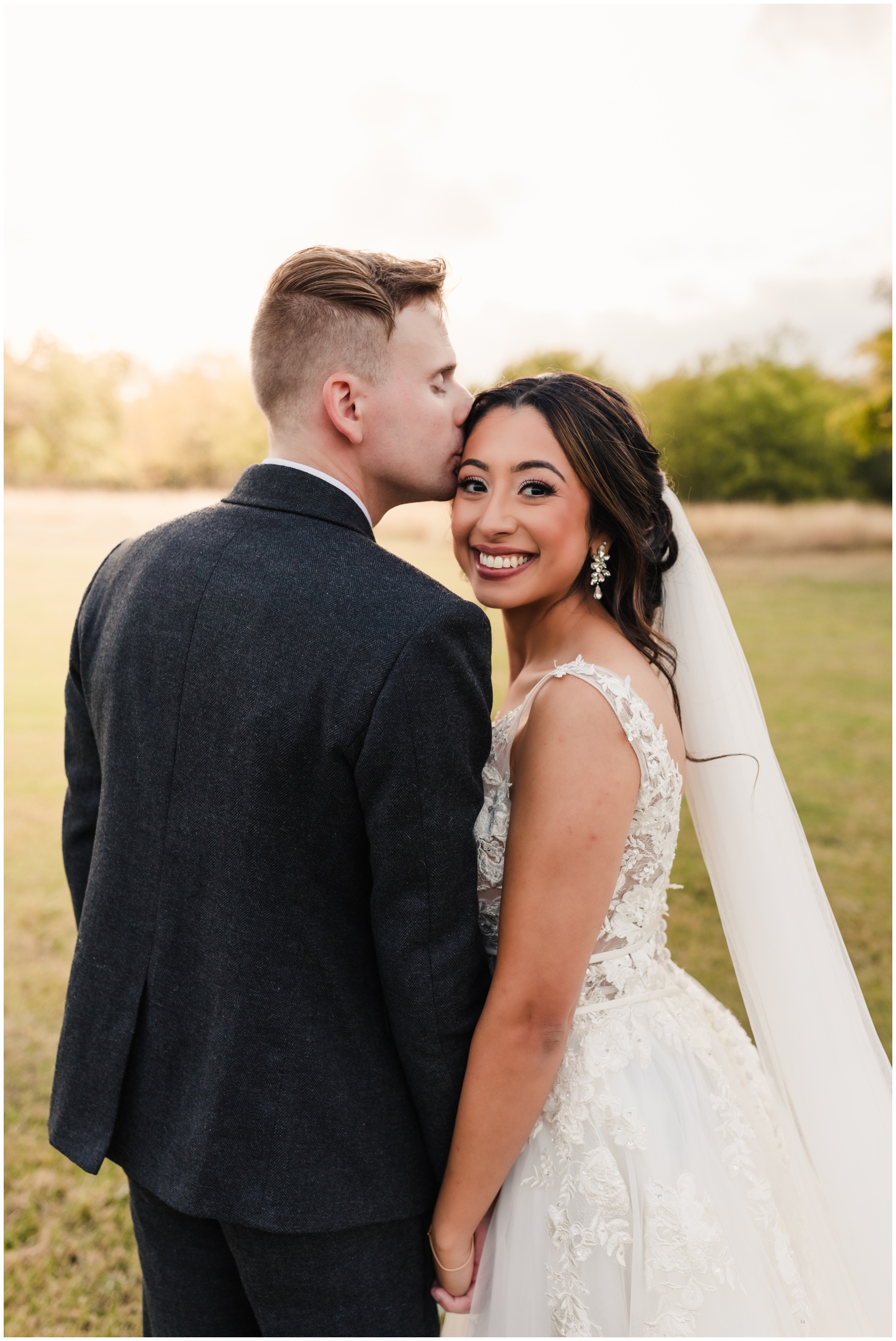 A bride smiles over her shoulder as her groom kisses her head in a field at sunset at The Emerson Wedding Venue