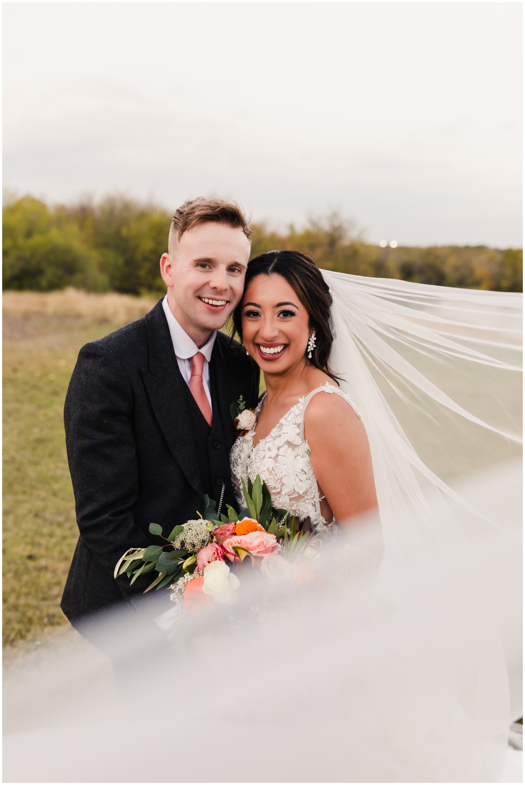 Happy newlyweds stand cheek to cheek in a windy field at sunset smiling big at The Emerson Wedding Venue