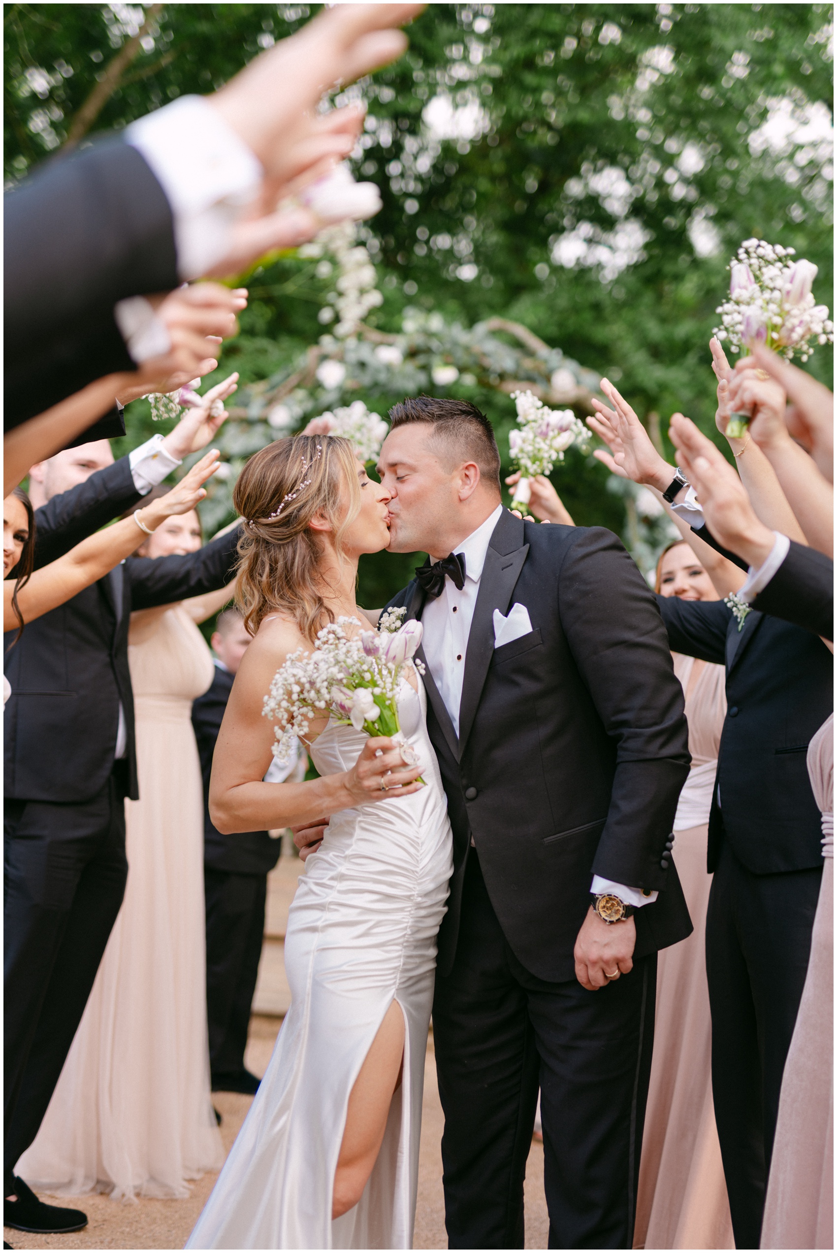 Newlyweds kiss while surrounded by their wedding party with hands up over them