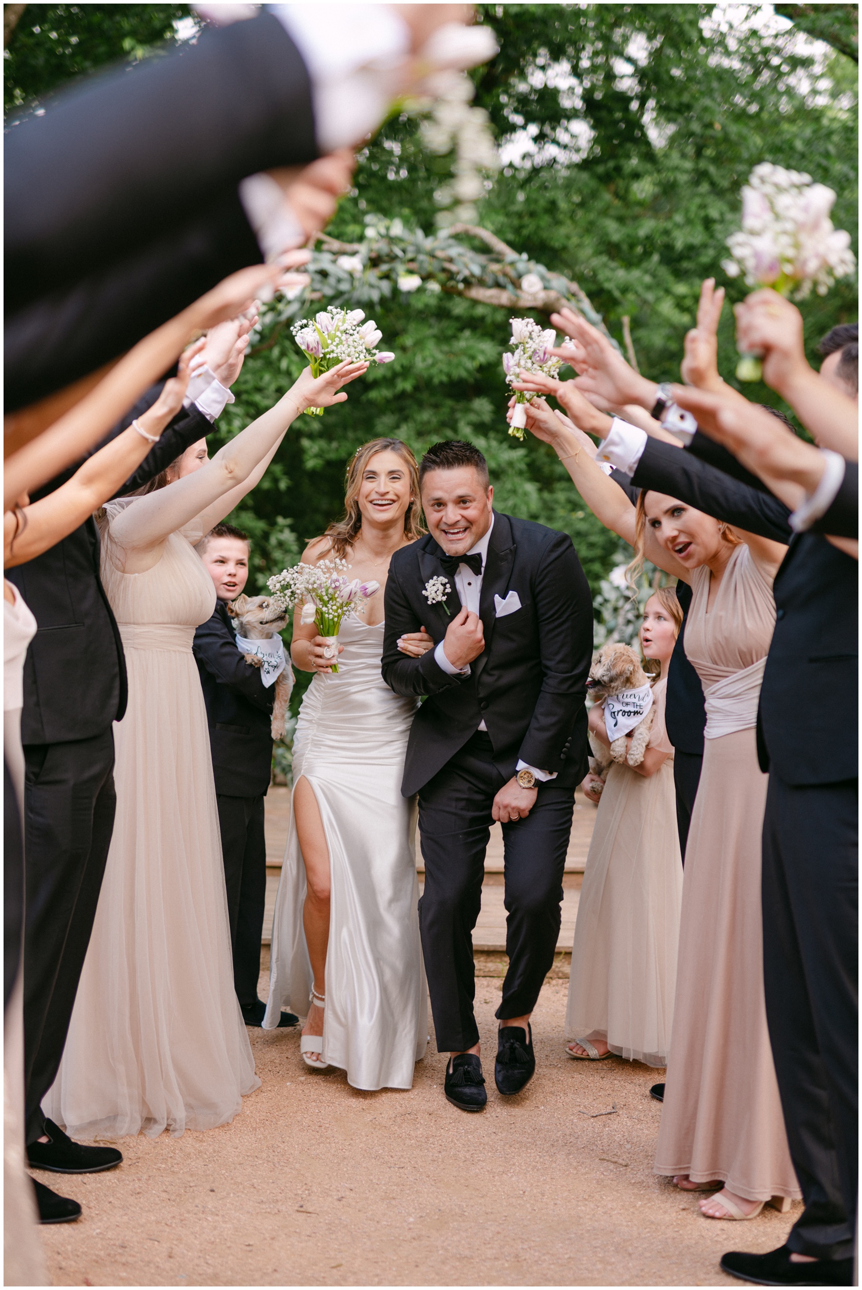 Newlyweds laugh while walking under an arch of hands from the wedding party at The French Farmhouse